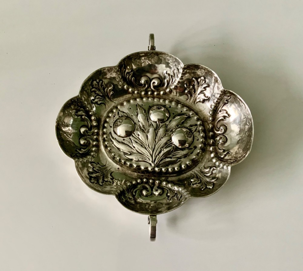 An oval German silver wine taster featuring repousse fruit central decoration, scalloped borders with acanthus scroll details, and loop handles.