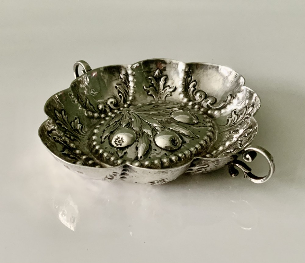 An oval German silver wine taster featuring repousse fruit central decoration, scalloped borders with acanthus scroll details, and loop handles. View III