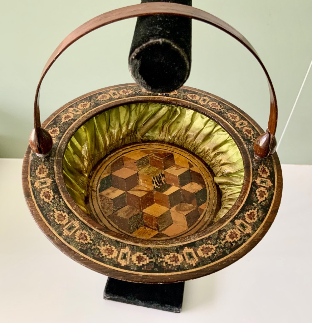 A circular Regency era wooden sewing basket featuring geometric Tunbridge Ware mosaic inlay along the rim, an optical cube parquetry base, green pleated silk interior lining, and an arched handle.