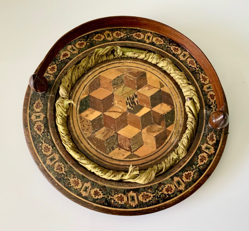 A circular Regency era wooden sewing basket featuring geometric Tunbridge Ware mosaic inlay along the rim, an optical cube parquetry base, green pleated silk interior lining, and an arched handle. View II