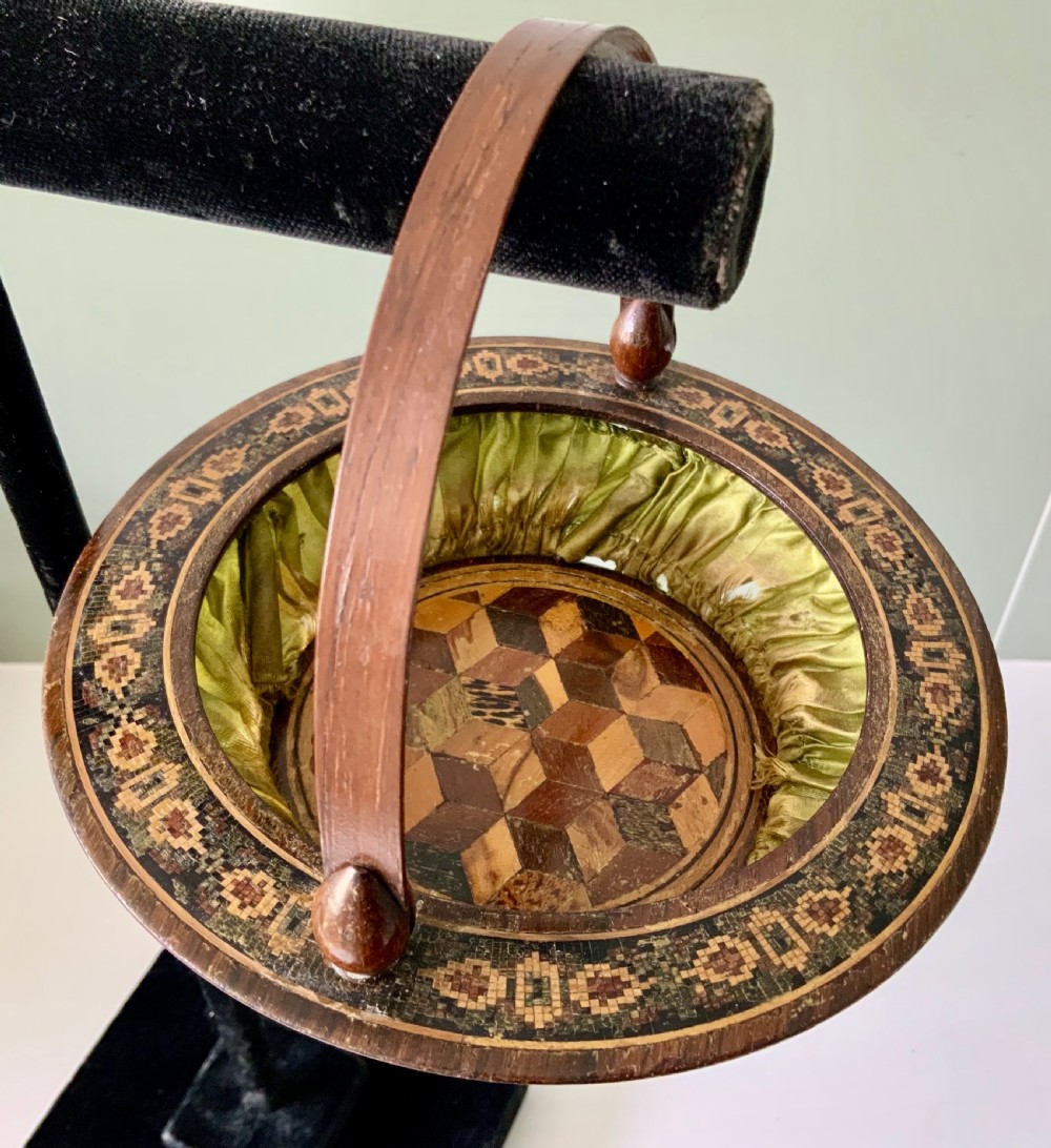 A circular Regency era wooden sewing basket featuring geometric Tunbridge Ware mosaic inlay along the rim, an optical cube parquetry base, green pleated silk interior lining, and an arched handle. View IV