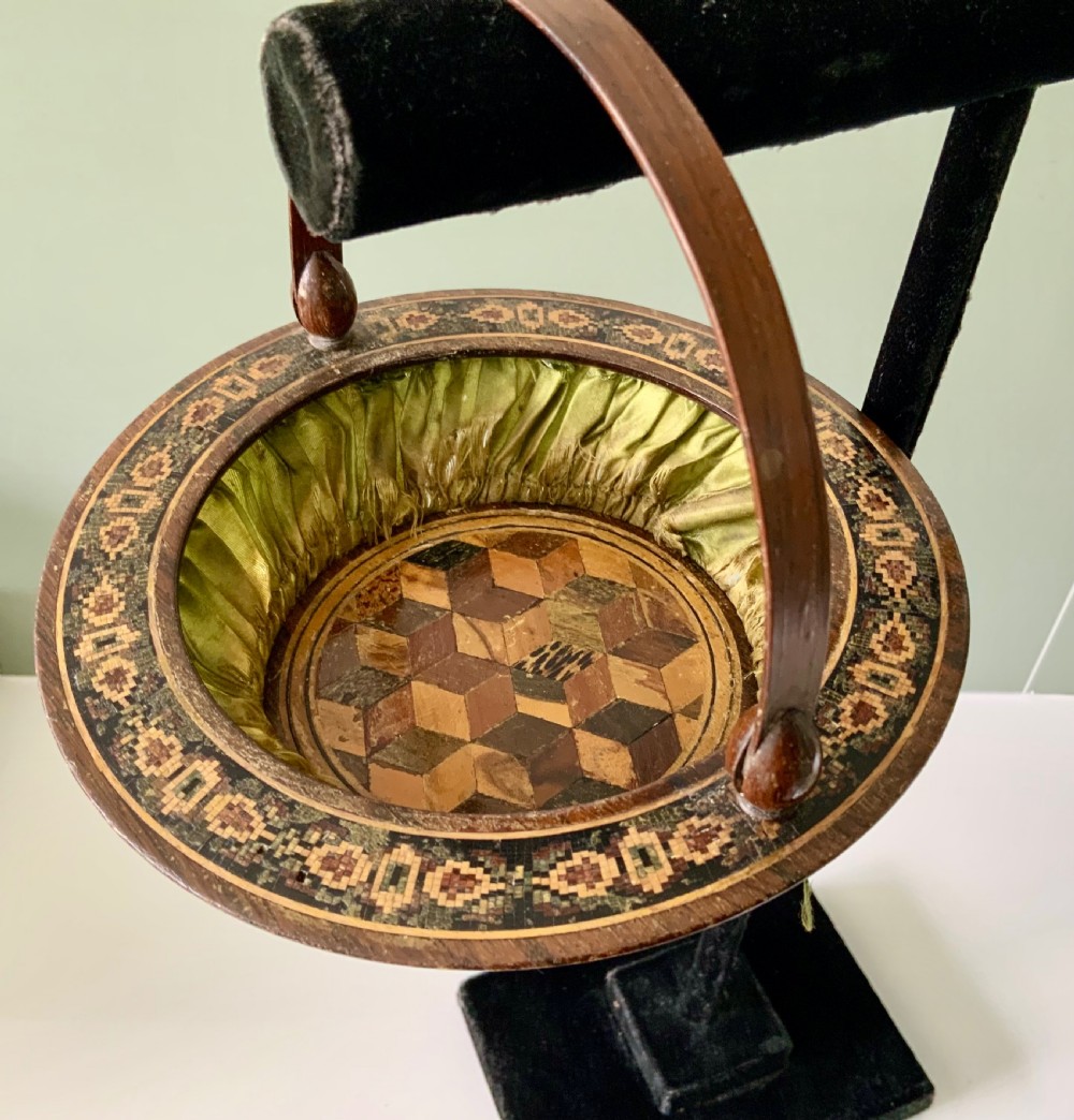 A circular Regency era wooden sewing basket featuring geometric Tunbridge Ware mosaic inlay along the rim, an optical cube parquetry base, green pleated silk interior lining, and an arched handle. View V