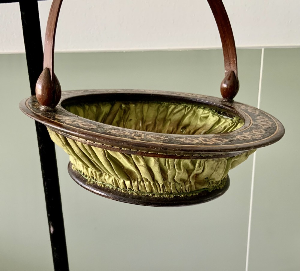 A circular Regency era wooden sewing basket featuring geometric Tunbridge Ware mosaic inlay along the rim, an optical cube parquetry base, green pleated silk interior lining, and an arched handle. View VI
