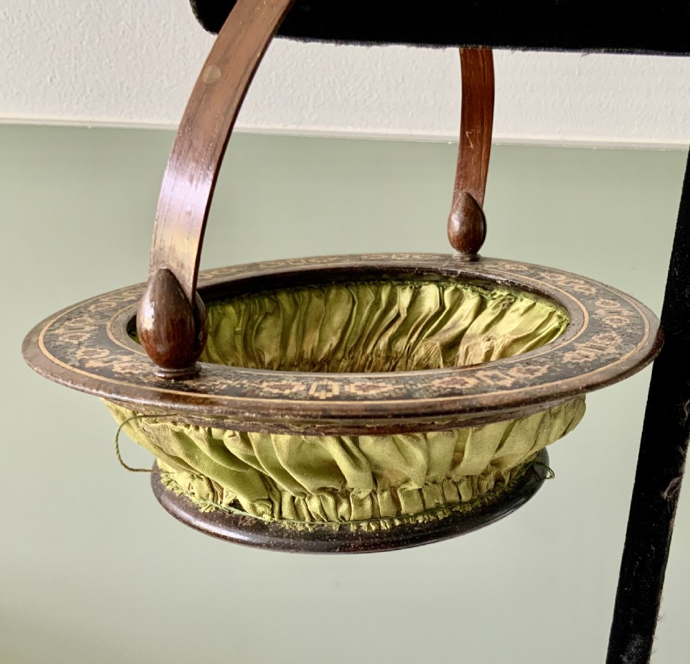 A circular Regency era wooden sewing basket featuring geometric Tunbridge Ware mosaic inlay along the rim, an optical cube parquetry base, green pleated silk interior lining, and an arched handle. View VII