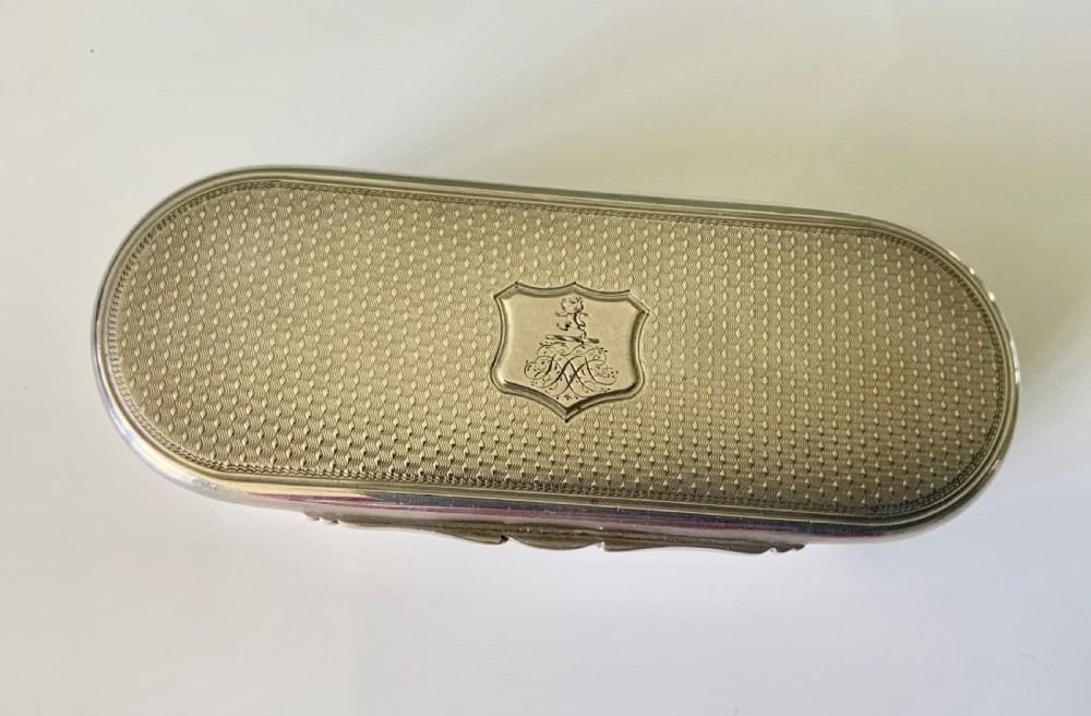 An elongated oval sterling silver snuff box featuring fine engine-turned surface pattern and a central engraved crest shield. View II