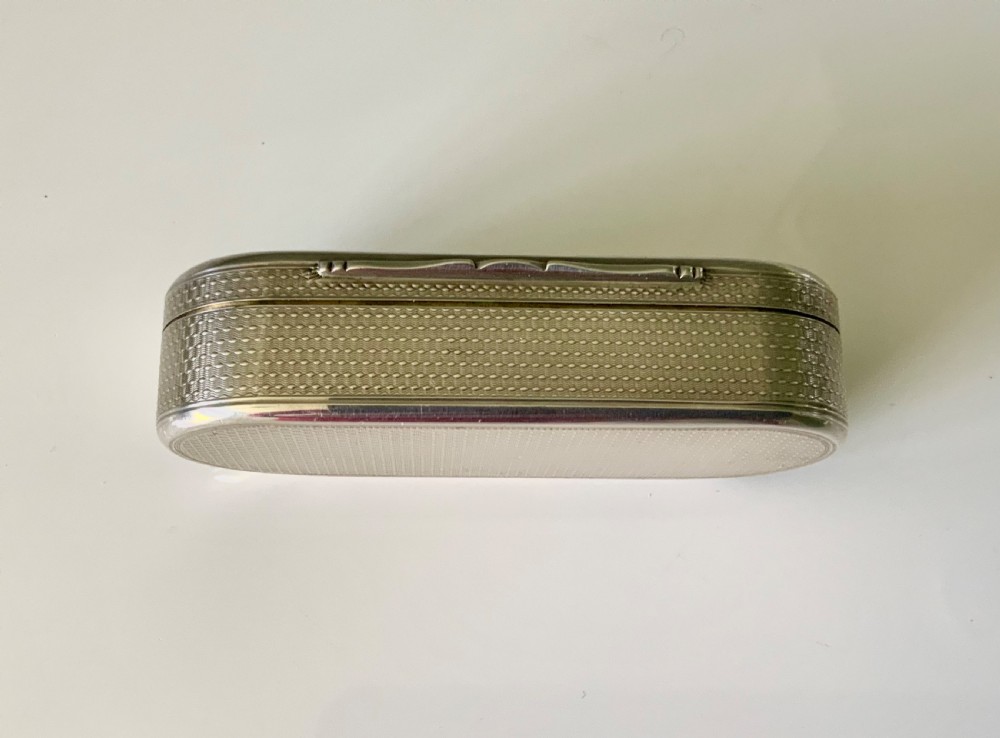 An elongated oval sterling silver snuff box featuring fine engine-turned surface pattern and a central engraved crest shield. View III