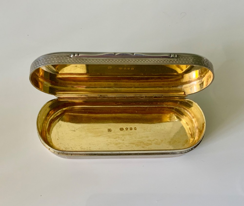 An elongated oval sterling silver snuff box featuring fine engine-turned surface pattern and a central engraved crest shield. View V