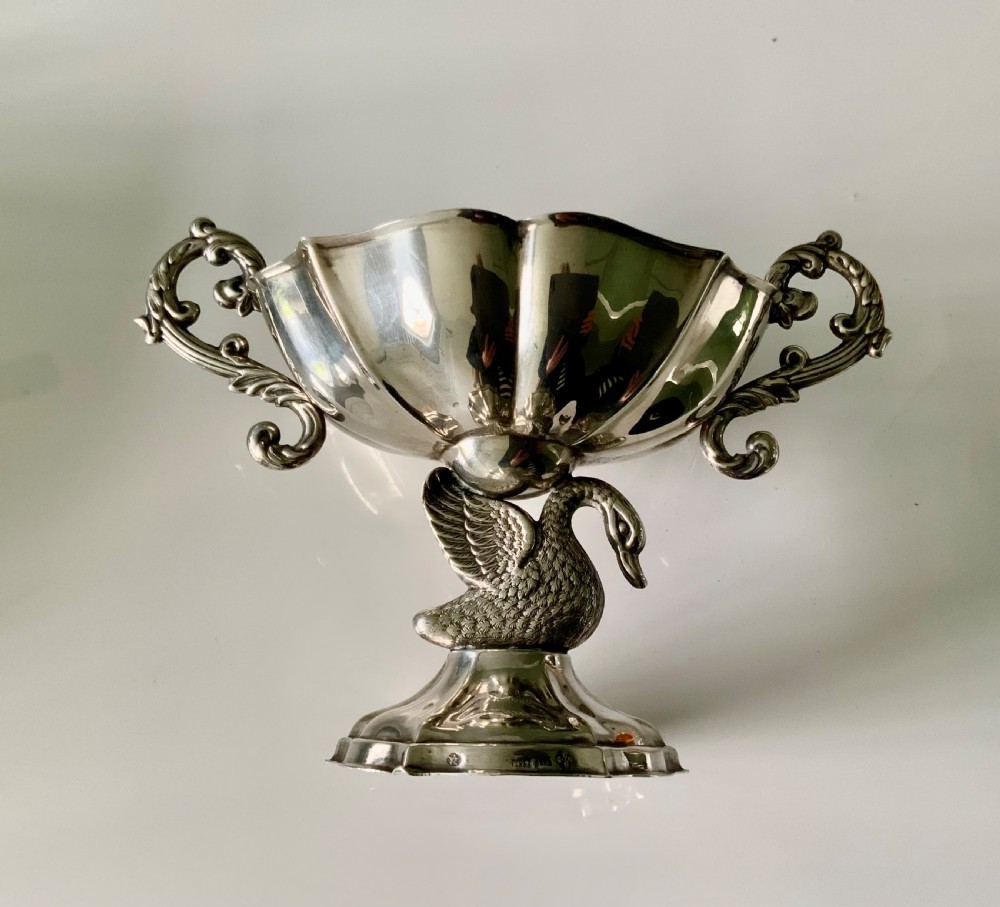 A polished silver dish featuring a lobed rim, two ornate scroll handles, and a central swan motif on a flared pedestal base.