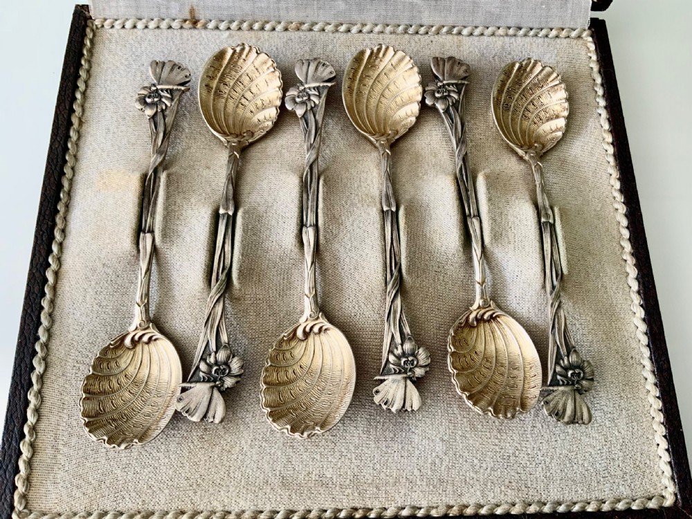 A set of six Art Nouveau silver teaspoons with shell bowls and floral stems, displayed inside an open fitted cloth-lined presentation box. View II