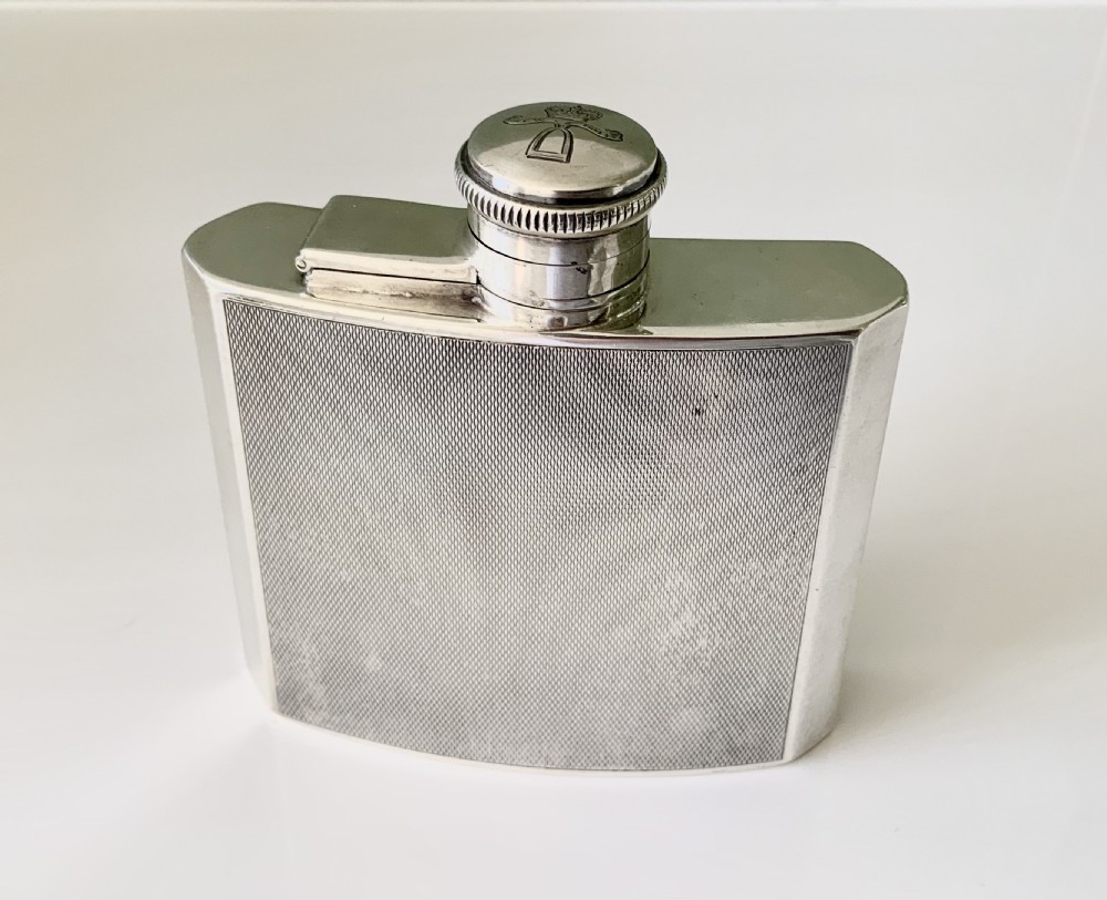 An Art Deco silver hip flask featuring a subtle engine-turned pattern, a contoured body, and a hinged bayonet cap engraved with an emblem.