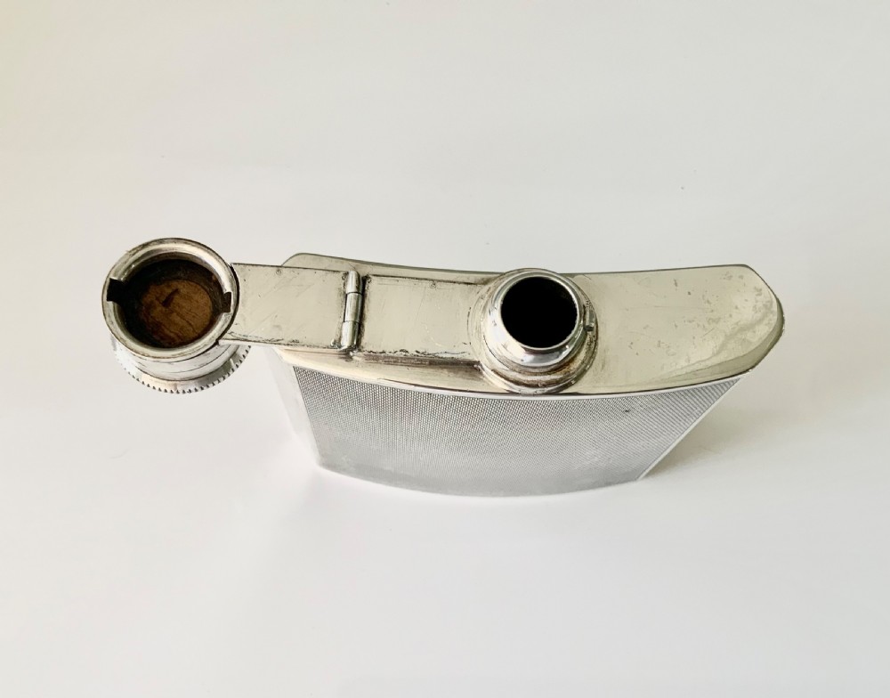 An Art Deco silver hip flask featuring a subtle engine-turned pattern, a contoured body, and a hinged bayonet cap engraved with an emblem. View IV