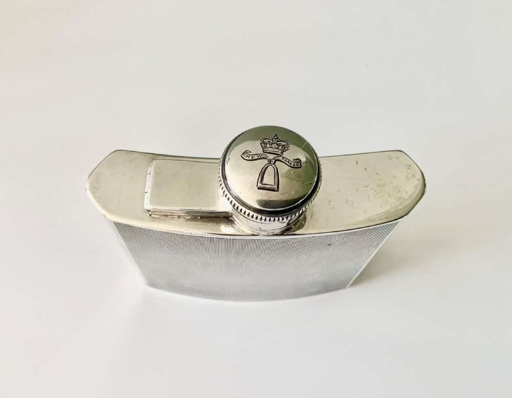 An Art Deco silver hip flask featuring a subtle engine-turned pattern, a contoured body, and a hinged bayonet cap engraved with an emblem. View V