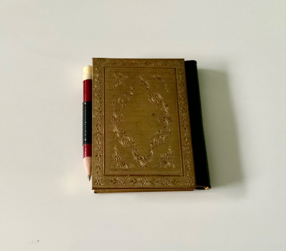 A late nineteenth-century miniature notebook featuring an embossed brass front cover with floral scrollwork, a dark leather spine, and an attached pencil. View II