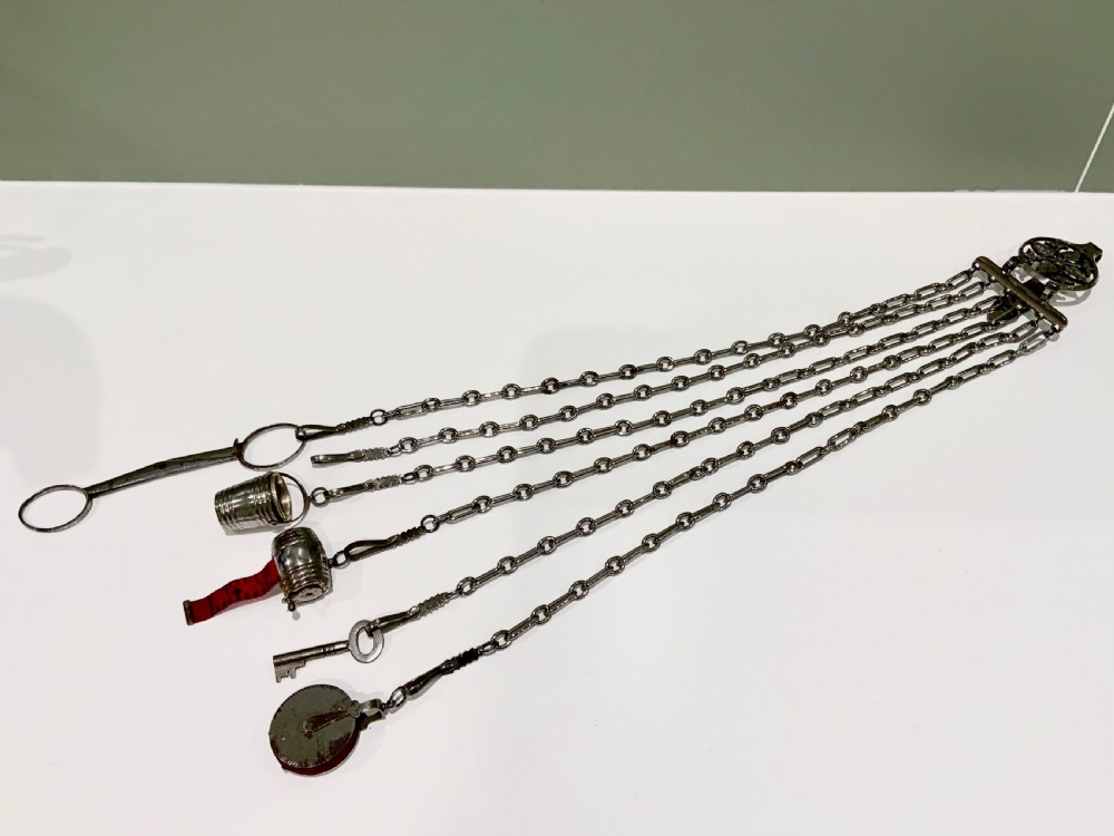 A nineteenth-century steel chatelaine featuring an ornamental waist clip and five suspended chains holding sewing tools, a key, a thimble, and a tape measure.