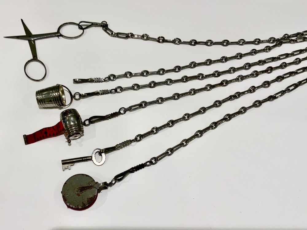 A nineteenth-century steel chatelaine featuring an ornamental waist clip and five suspended chains holding sewing tools, a key, a thimble, and a tape measure. View V