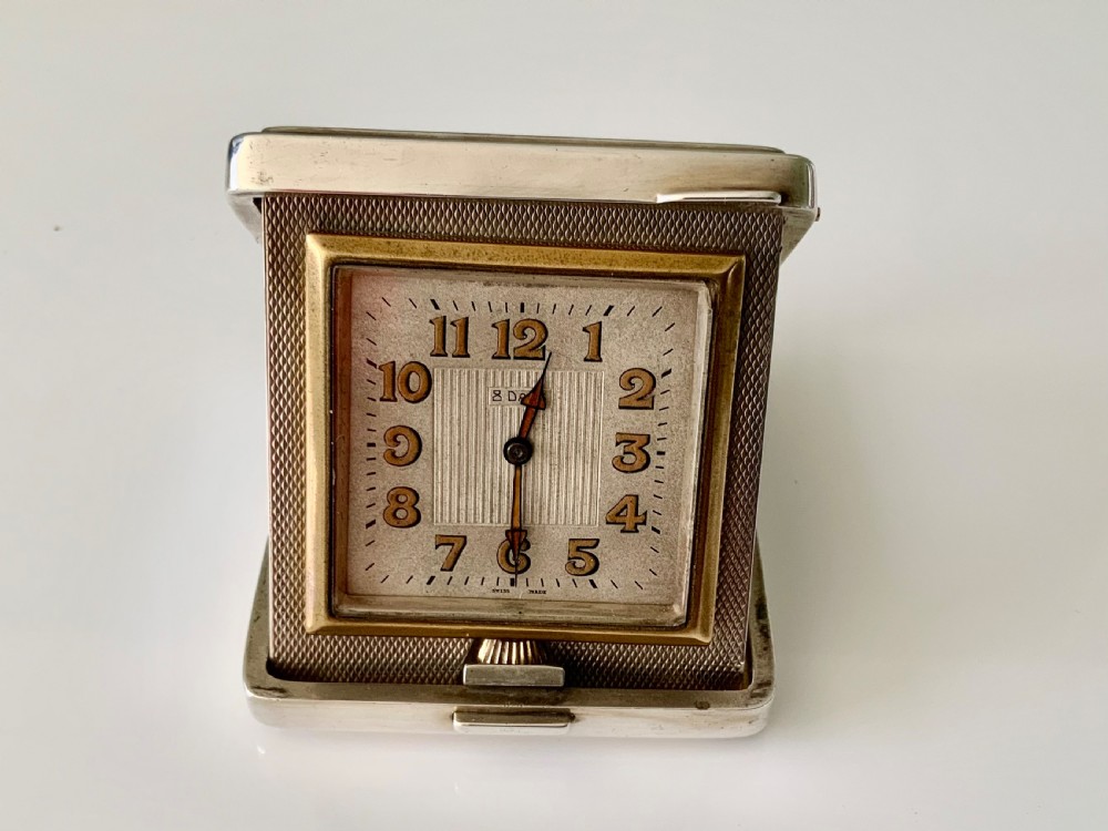 A square Art Deco silver travel clock featuring an engine-turned hinged case, brass bezel, and cream dial with gold Arabic numerals.
