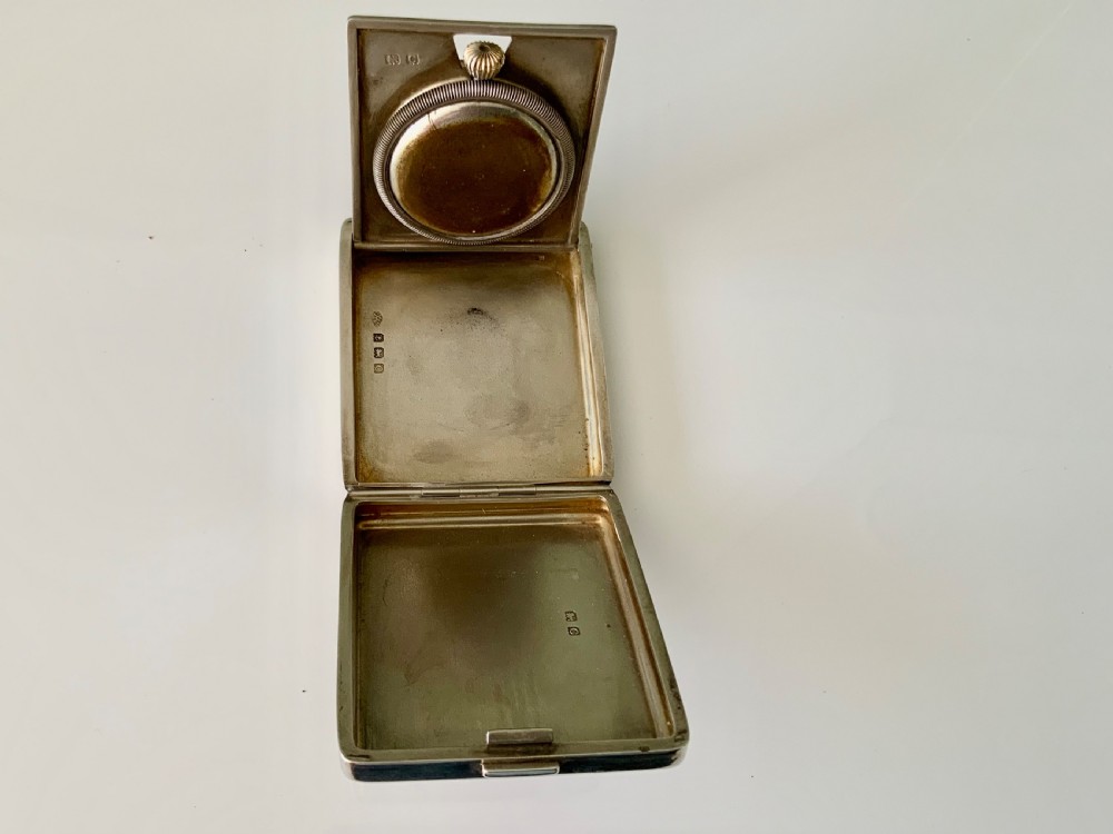A square Art Deco silver travel clock featuring an engine-turned hinged case, brass bezel, and cream dial with gold Arabic numerals. View III