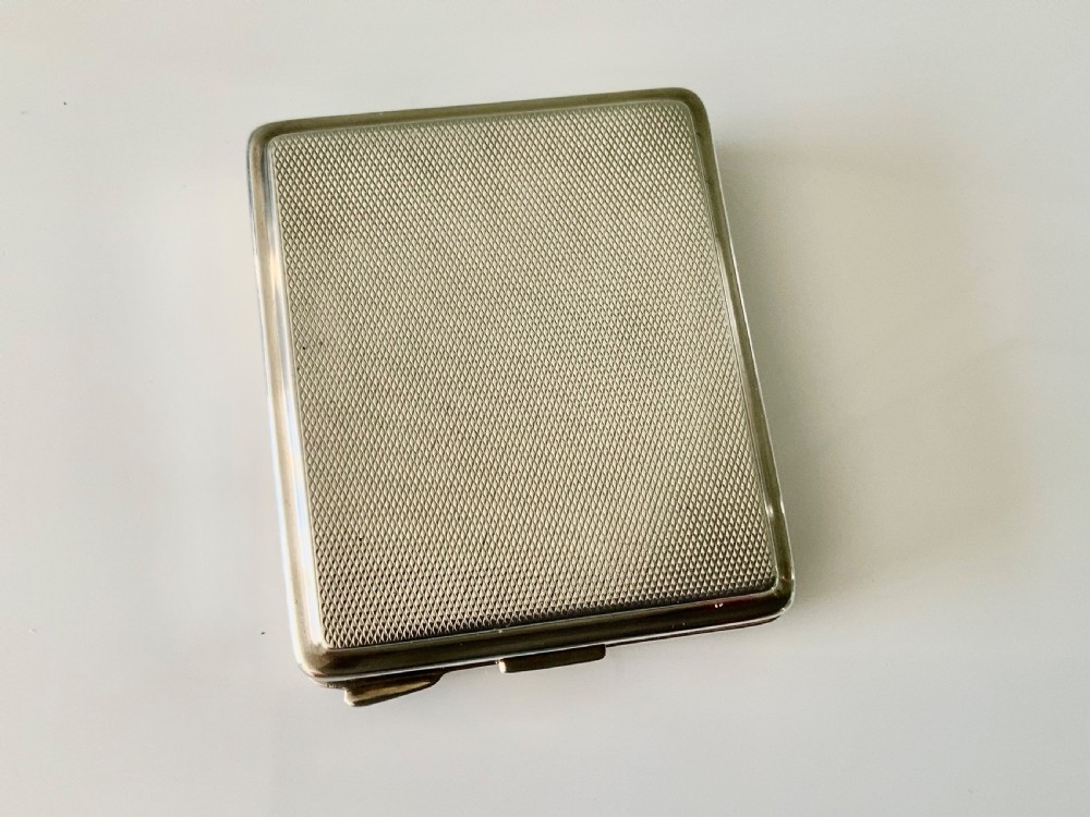 A square Art Deco silver travel clock featuring an engine-turned hinged case, brass bezel, and cream dial with gold Arabic numerals. View V