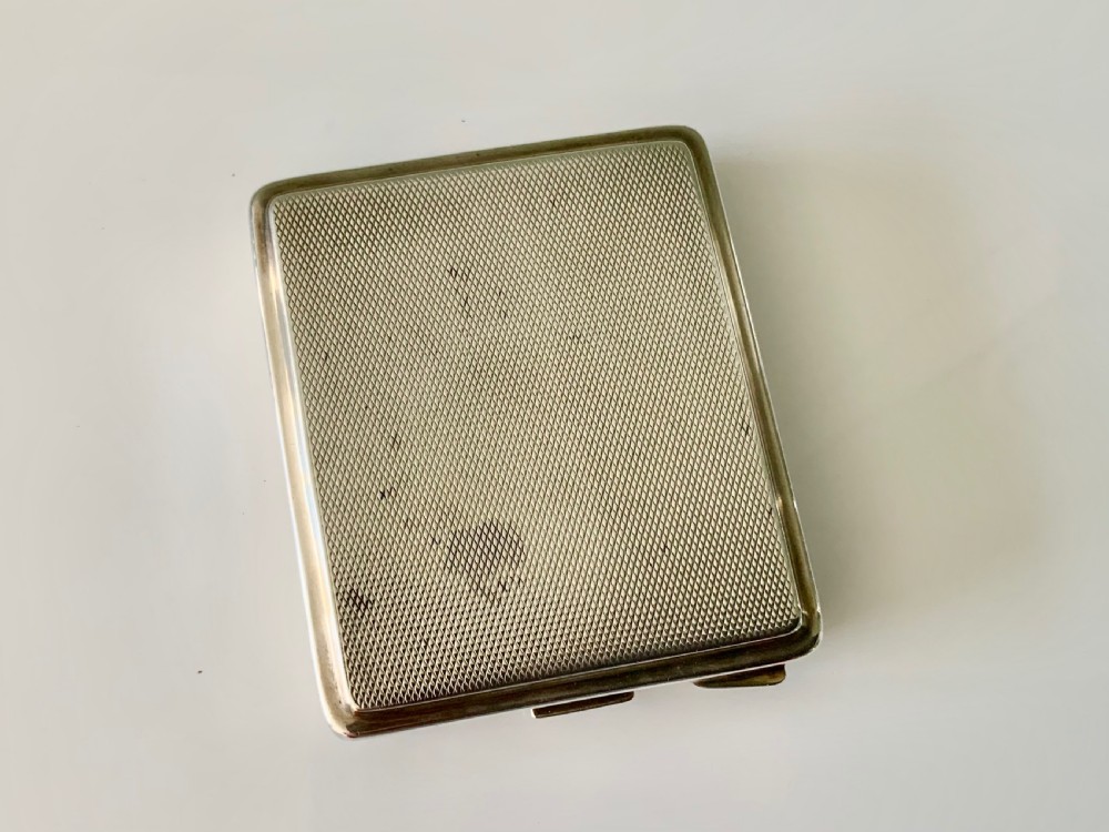 A square Art Deco silver travel clock featuring an engine-turned hinged case, brass bezel, and cream dial with gold Arabic numerals. View VI