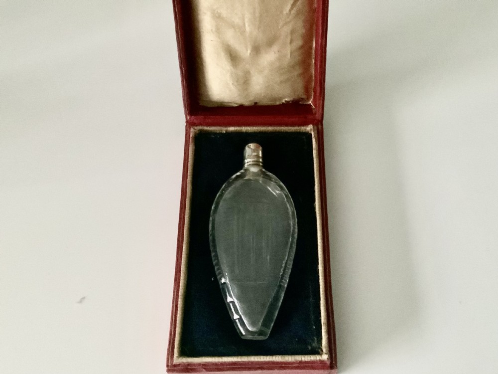 A teardrop-shaped clear glass scent bottle with a silver cap resting inside an open red leather plush-lined case.