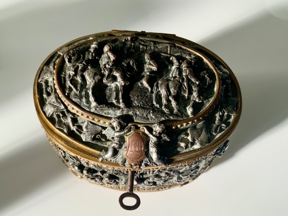 An oval antique French metal casket featuring relief panels of figures on horseback and cherubs holding an escutcheon with a key.