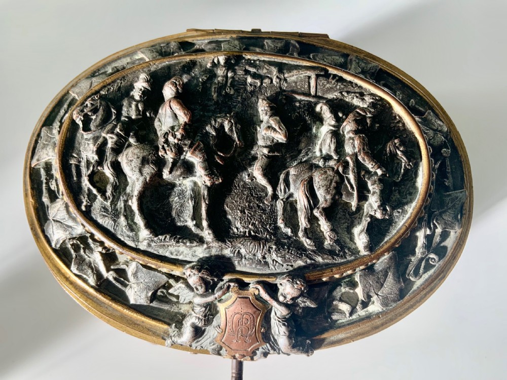 An oval antique French metal casket featuring relief panels of figures on horseback and cherubs holding an escutcheon with a key. View II