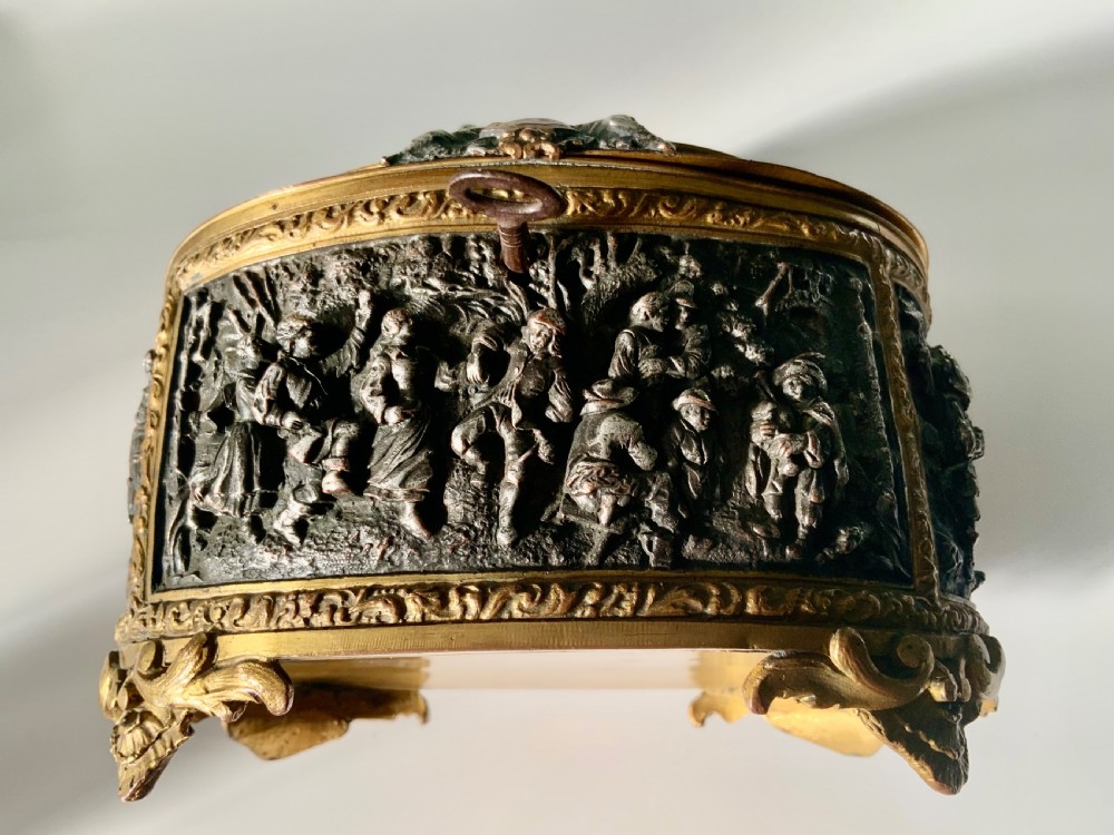 An oval antique French metal casket featuring relief panels of figures on horseback and cherubs holding an escutcheon with a key. View III