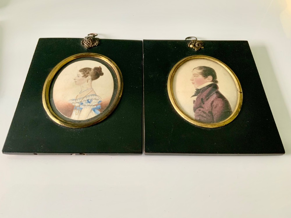 A pair of early nineteenth-century watercolour portrait miniatures depicting a lady and a gentleman, mounted in oval brass bezels set within rectangular black ebonised frames with top hanging loops.