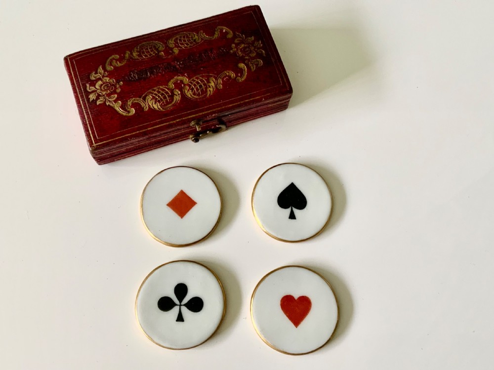 A set of four round white porcelain whist counters featuring suit motifs alongside a red gilt-tooled leather storage box.