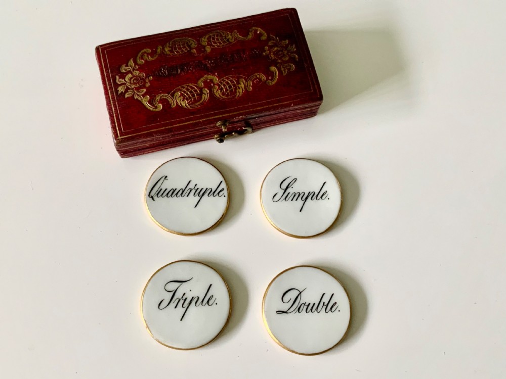 A set of four round white porcelain whist counters featuring suit motifs alongside a red gilt-tooled leather storage box. View II