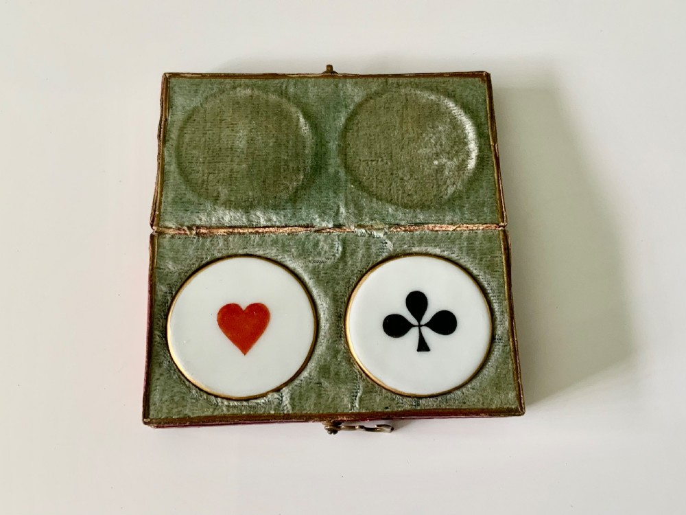 A set of four round white porcelain whist counters featuring suit motifs alongside a red gilt-tooled leather storage box. View IV