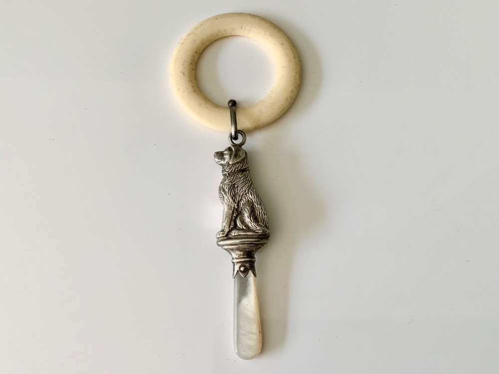 An antique silver baby rattle shaped like a seated dog with a mother of pearl handle, attached to a cream-coloured teething ring.