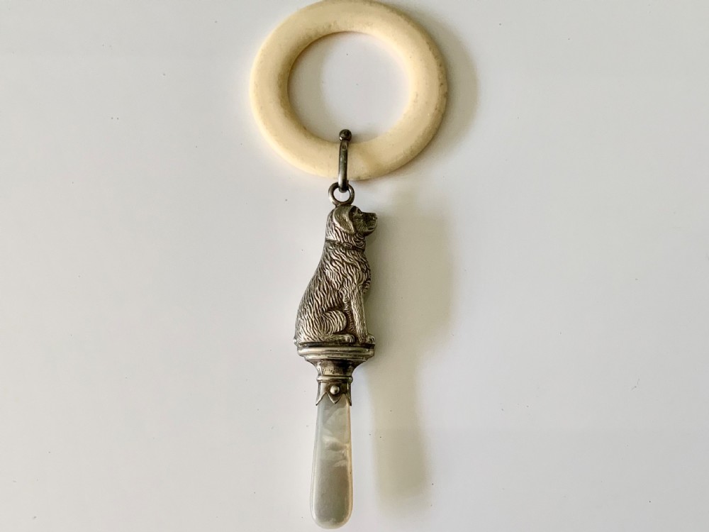 An antique silver baby rattle shaped like a seated dog with a mother of pearl handle, attached to a cream-coloured teething ring. View II