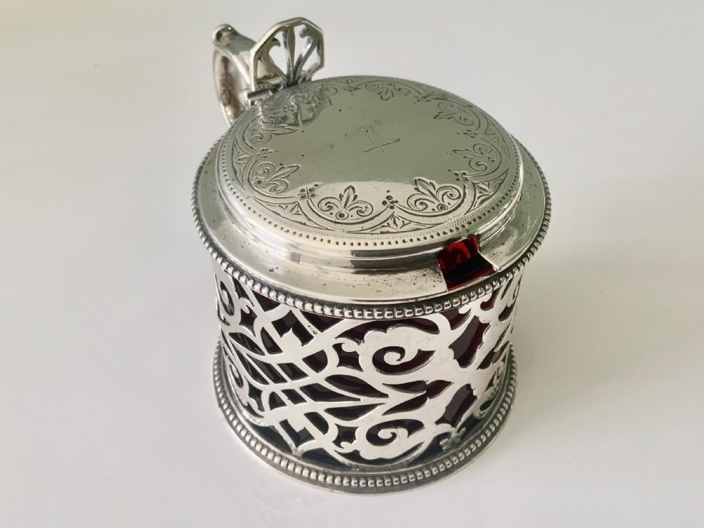 A pierced silver mustard pot with an engraved hinged lid, beaded rims, and a visible ruby red glass liner.