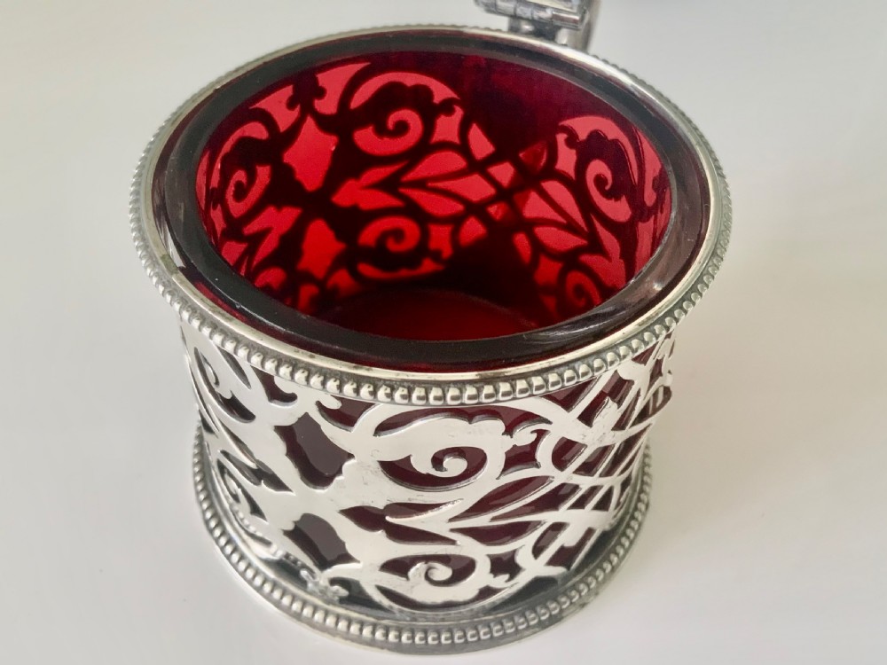 A pierced silver mustard pot with an engraved hinged lid, beaded rims, and a visible ruby red glass liner. View VI