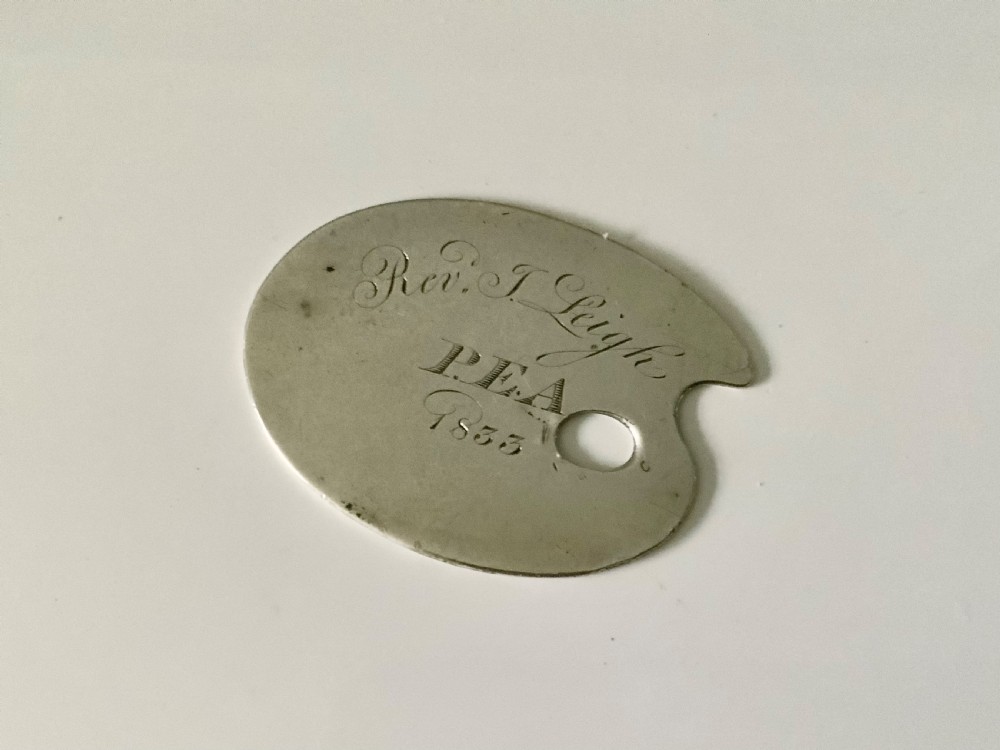 An engraved silver token shaped like an artists palette with a thumb hole, featuring cursive text that reads Rev. J. Leigh P.E.A. 1833.