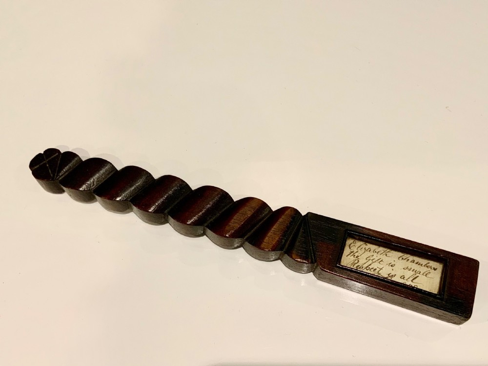 A dark carved wooden 19th-century knitting sheath featuring a heavy spiral-twisted shaft and an inset glass-covered handwritten inscription near the base.