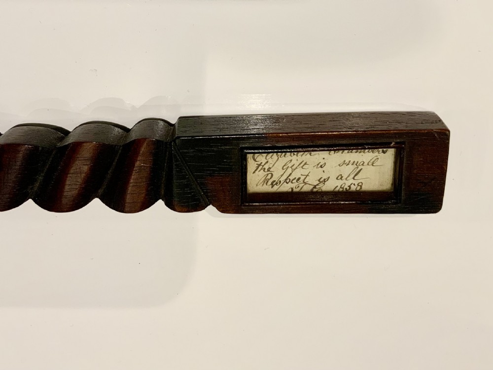 A dark carved wooden 19th-century knitting sheath featuring a heavy spiral-twisted shaft and an inset glass-covered handwritten inscription near the base. View II