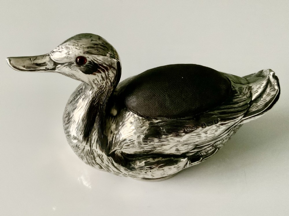 An Edwardian silver novelty pin cushion shaped like a resting duck, featuring detailed feather engraving, red glass eyes, and a dark velvet cushion on its back.