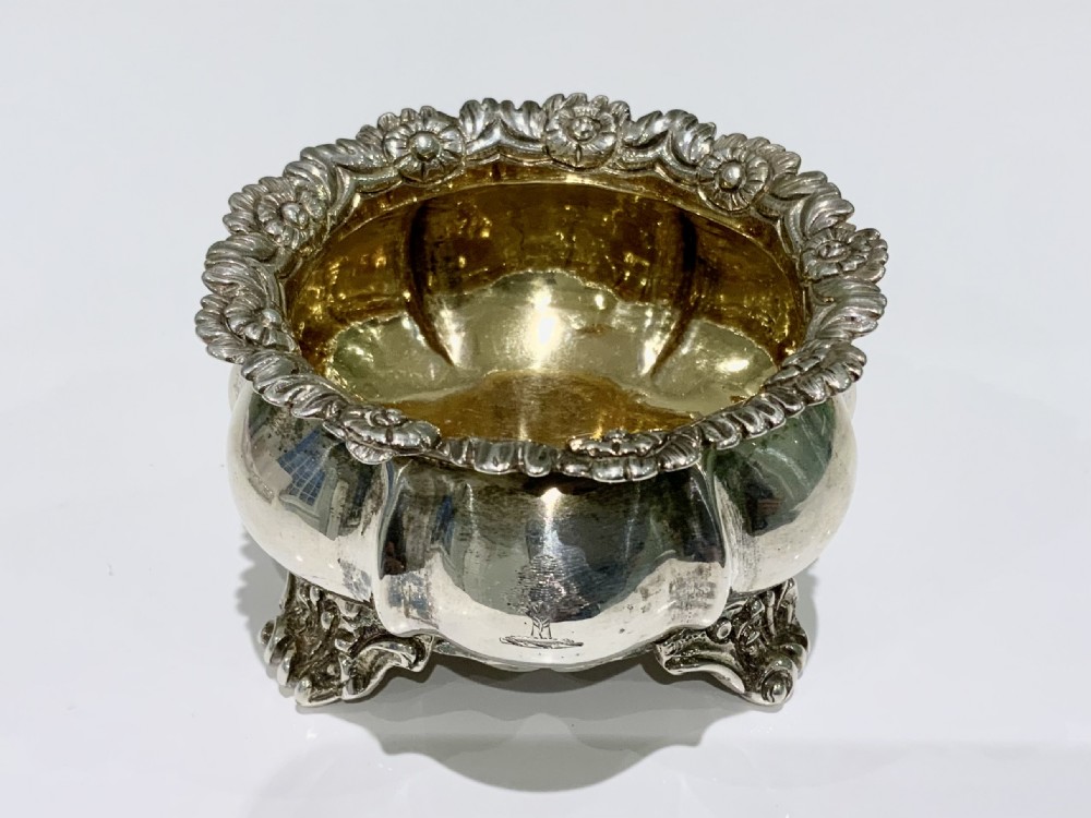 A round Georgian sterling silver open salt cellar featuring a gilded interior, a heavily embossed floral rim, a fluted body, and four cast scroll feet.