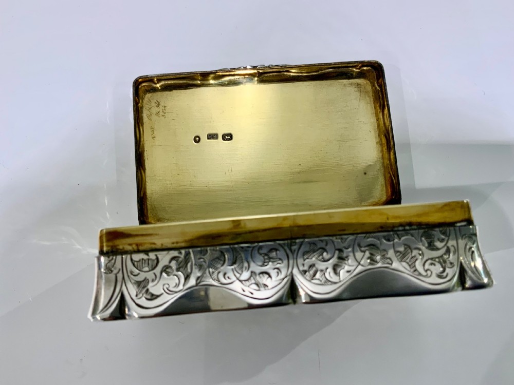An ornate rectangular silver snuff box with intricate scroll engraving and a central inscription on the hinged lid. View V