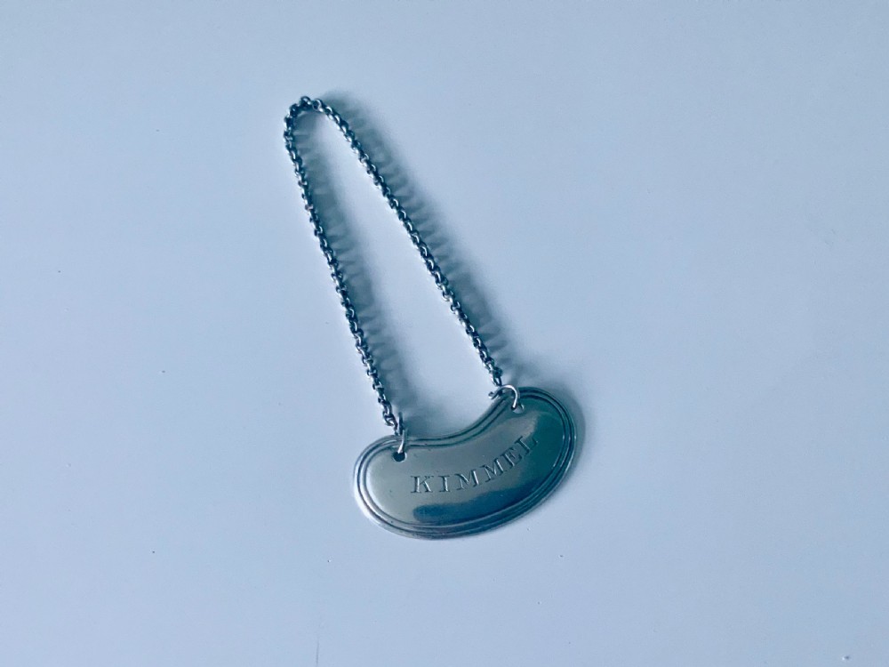A crescent-shaped silver decanter label engraved with the word KIMMEL suspended from a silver chain.