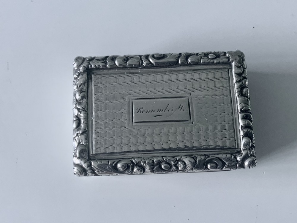 A small rectangular Georgian sterling silver vinaigrette featuring an engraved central cartouche with the inscription Remember Me, framed by basketweave engine turning and a floral cast border.