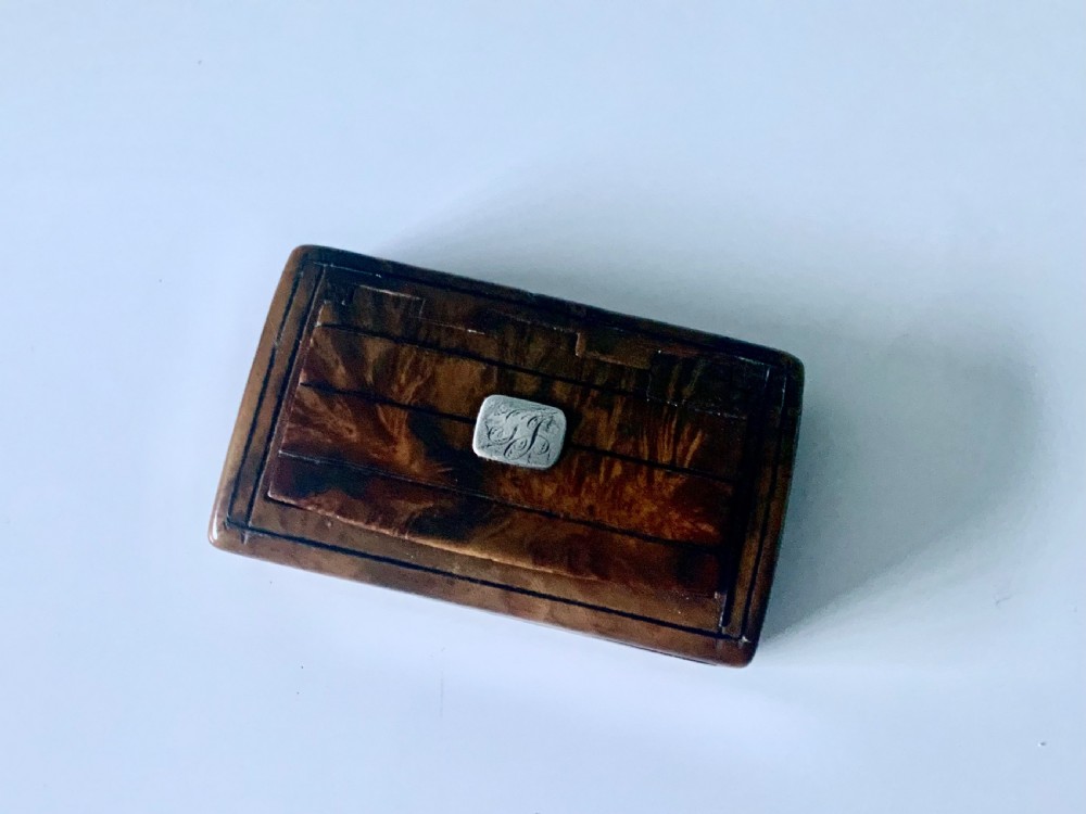 An overhead view of a rectangular antique burr wood snuff box featuring a distinctive swirling wood grain and a small engraved metal plaque set into the lid.