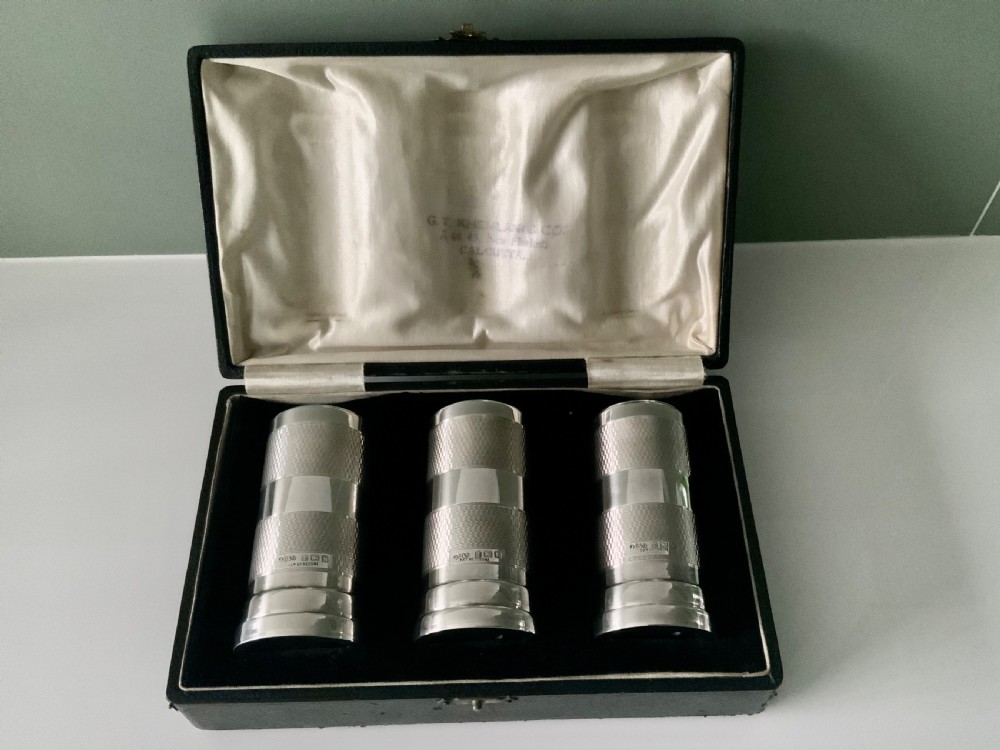 An open black fitted case displaying three cylindrical Art Deco sterling silver canisters with engine-turned decoration and satin lining.