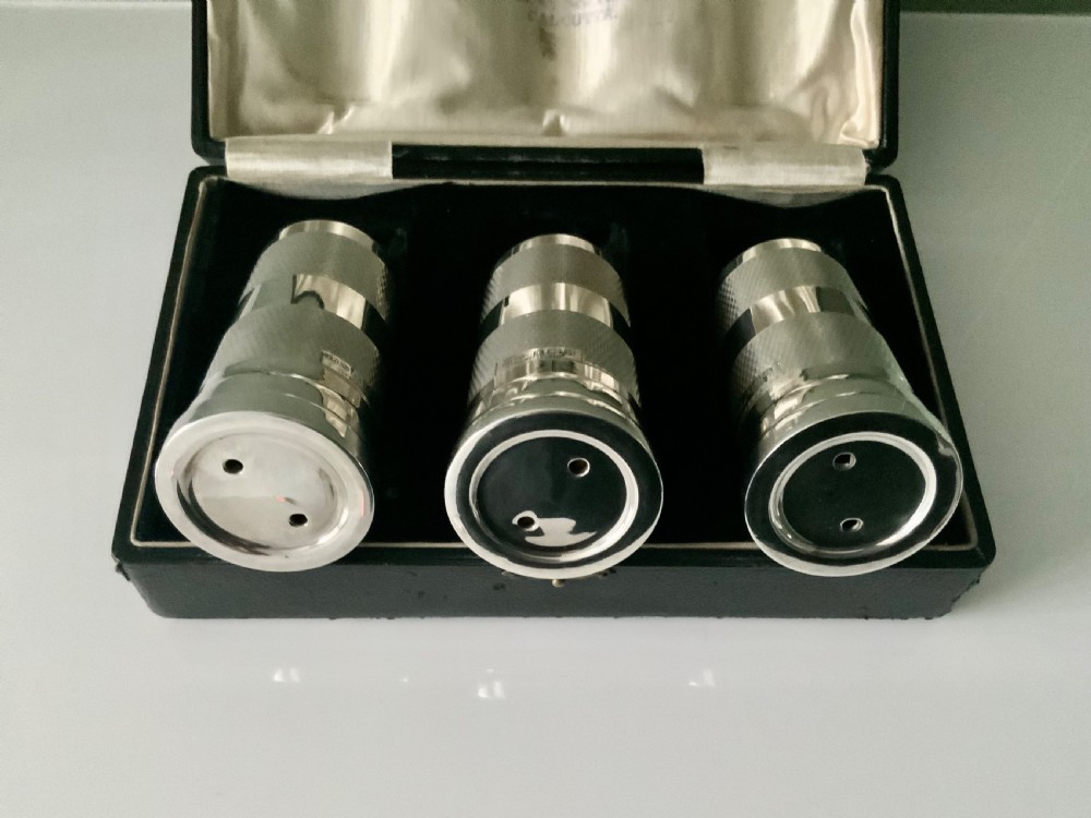 An open black fitted case displaying three cylindrical Art Deco sterling silver canisters with engine-turned decoration and satin lining. View III