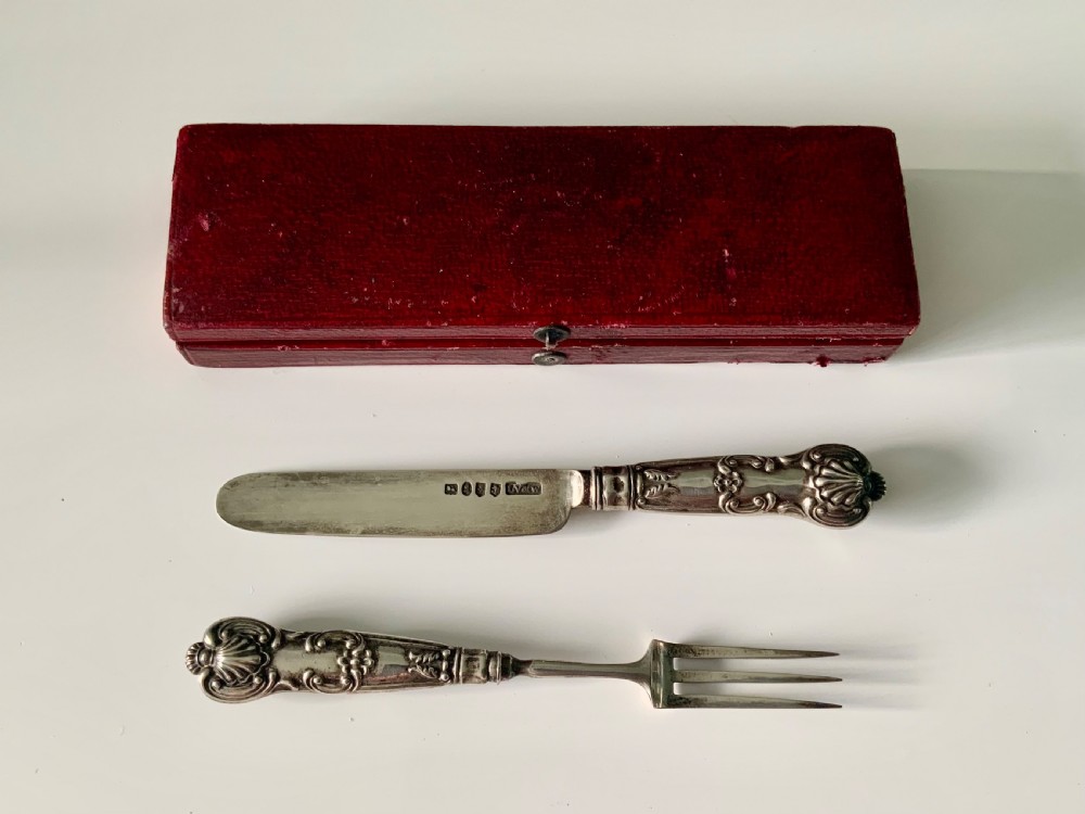 A boxed Georgian sterling silver childs knife and three-tined fork with shell motifs on the handles, displayed below a red fitted presentation case.