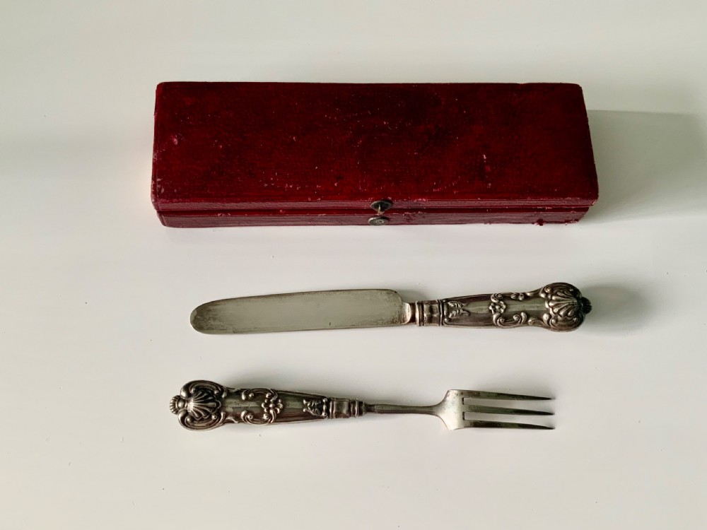 A boxed Georgian sterling silver childs knife and three-tined fork with shell motifs on the handles, displayed below a red fitted presentation case. View II