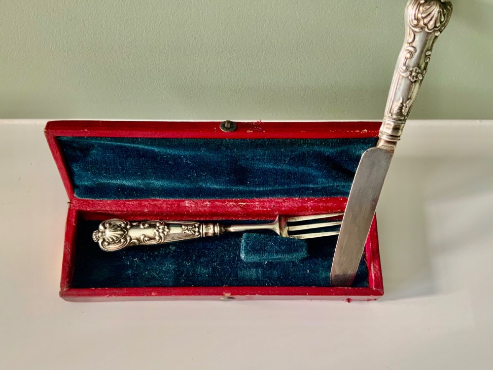 A boxed Georgian sterling silver childs knife and three-tined fork with shell motifs on the handles, displayed below a red fitted presentation case. View III