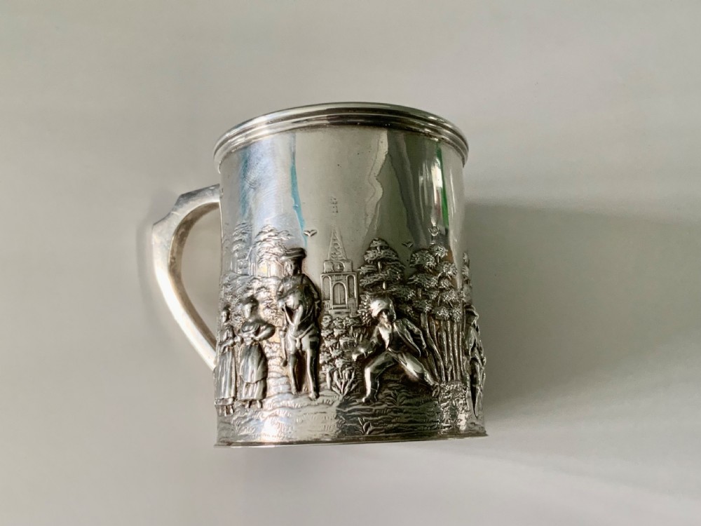 A small sterling silver christening mug decorated with embossed pastoral figures and a church landscape, featuring an angular side handle.
